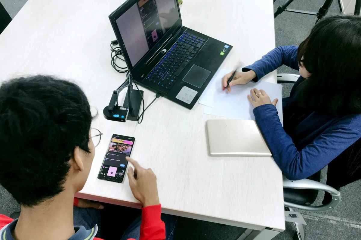 Two people testing a mobile interface at a table, with one taking notes beside a laptop.