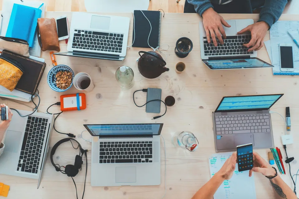 Overhead view of a shared wooden desk with four open laptops, people typing and using smartphones, notebooks and pens scattered around, a teapot and cups, external hard drives, headphones, and snacks, suggesting a collaborative work session.